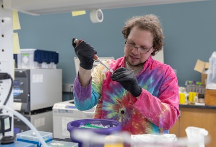 Man in tie-died lab coat and black latex gloves injects liquid into a test tube