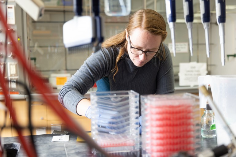 Woman with long reddish hair and glasses leans over a lab bench stacked with red plastic containers