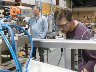 A man peers down at a piece of machinery with coil springs and blue tubingin an industrial setting. In the background a second man looks at something off camera.