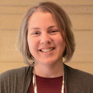 Headshot of Mari Magler, a white woman with short brown hair, smiling and wearing a red shirt and grey sweater