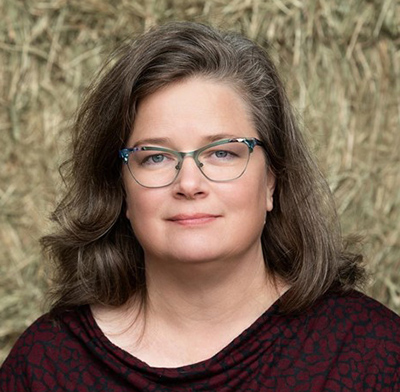 Portrait of Sarah Lloyd. She is a white woman with dark hair and glasses on. She is wearing a black and maroon shirt with a textured background behind her.