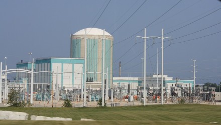 Large green and white silo-shaped building surrounded by power lines and other concrete buildings with blue sky in the background