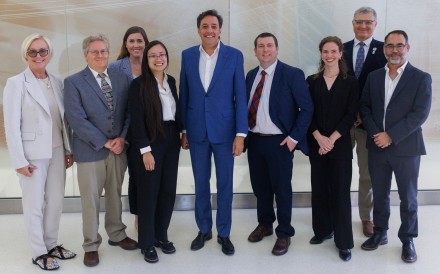 Nine people in business attire pose smiling for a group photo in a bright hallway, with a tall man in a bright blue suit standing at the center.