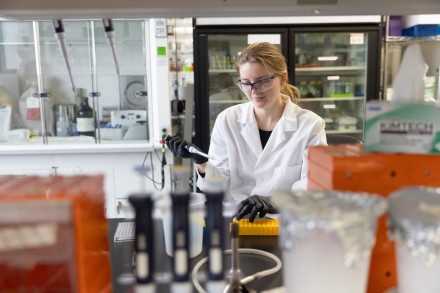 A woman in a white lab coat and lab glasses holds a pipette