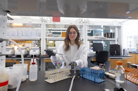 A woman in a white lab coat and latex gloves holds a flask of clear liquid in a laboratory