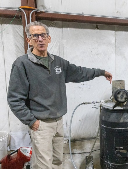 A man wearing a gray sweater and black-rimmed glasses looks at the camera while pointing to a black tank with hoses protruding from it and a piece of machinery on top