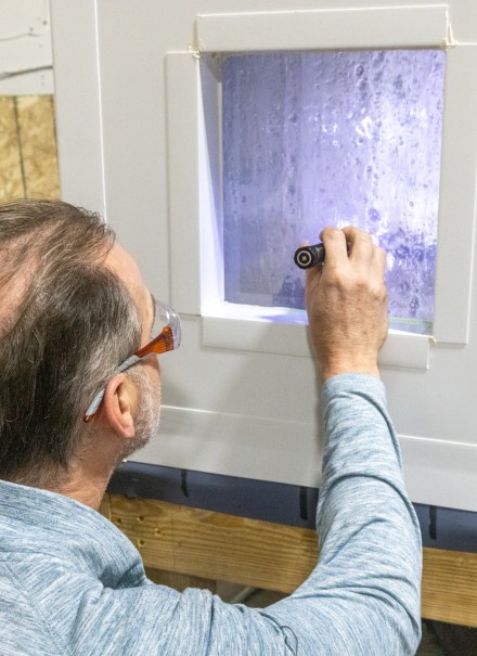 A man photographed from behind peers into a window in a piece of machinery while shinging a flashlight into the window. Clear liquid flows down the inside of the window.