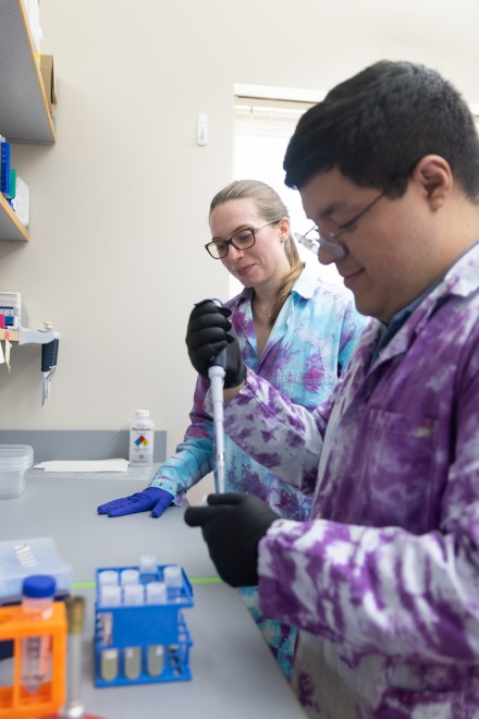 A man in a tie-died lab coat pipettes liquid as a woman observes