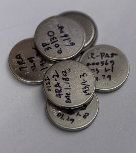 A pile of lithium-ion coin cell batteries hand-labeled in marker with sample identifiers and experimental parameters on a white surface.