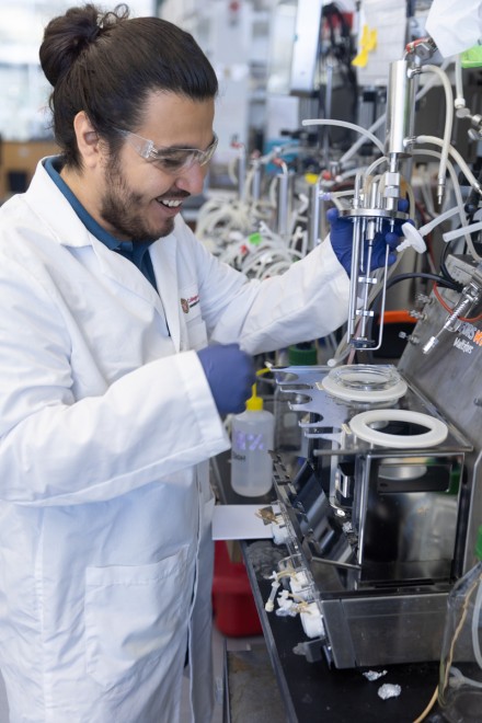 A man in a white lab coat and blue nitrile gloves inserts a piece of metal and glass equipment into a larger vessel at a black lab bench.