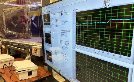 A wind tunnel test setup showing a real-time LabVIEW data acquisition display with windspeed (10.9 m/s), voltage, current, and power output readings, with the wind tunnel and a small turbine model visible in the background.