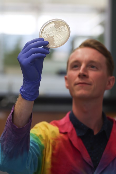 A white man in a pink, yellow and blue lab coat looks up at the Petri dish he holds in a blue gloved hand