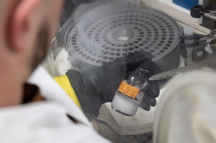 Closeup of a hand in a black rubber glove holding a glass container and a pipette