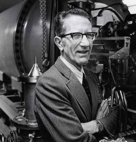 Man in suit jacket and tie with horned-rim glasses standing with arms crossed in front of machinery