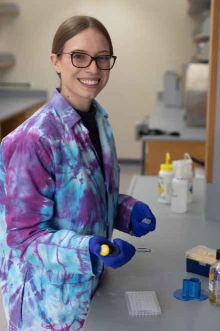 Woman in a purple tie-dyed lab coat stands at a lab bench holding a pipette and smiling towards the camera