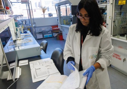 A researcher in a lab coat and blue gloves reviews a lab notebook at a benchtop, with analytical instruments and a fume hood visible in the background.