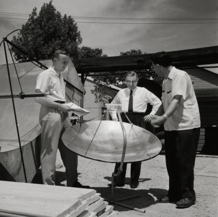 Black and white photo of three men standing around a large metal dish pointed upward.