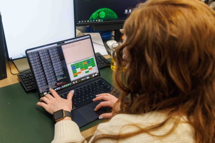 A researcher with long red hair, seen from behind, types on a laptop displaying terminal windows and a data heatmap, with a microscopy image on a second monitor.