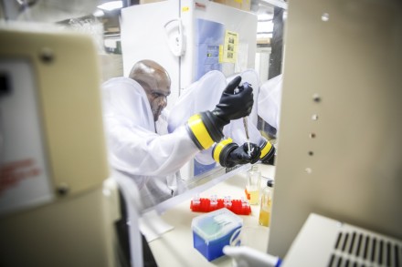 Man in white lab coat uses rubber sleeves to reach through a plastic barier