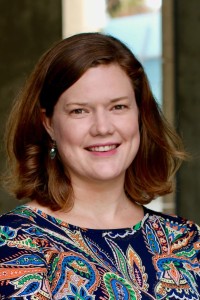 A woman with shoulder length brown hair wearing a blue, orange and green paisley shirt