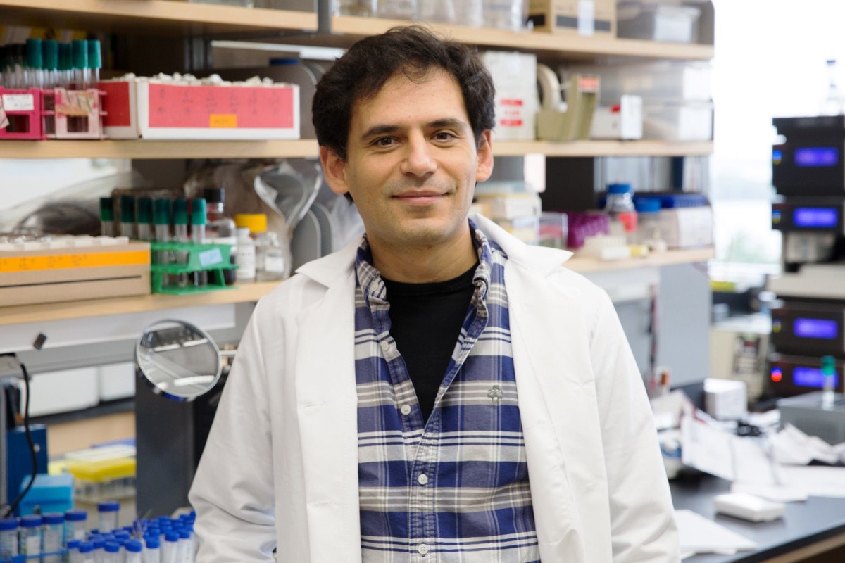 Man in blue checked shirt and white lab coat standing in front of a lab bench