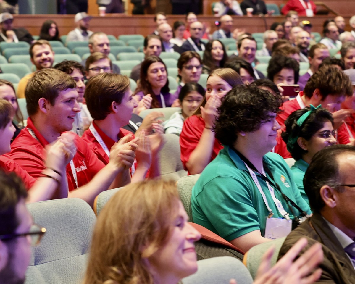 Students in red shirts seated in auditorium clap as woman in red shirt covers her mouth with her hand in surprise