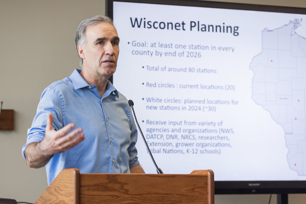 Man in blue shirt stands behind a podium gesturing with one hand. A screen behind him is titled "Wisconet Planning"