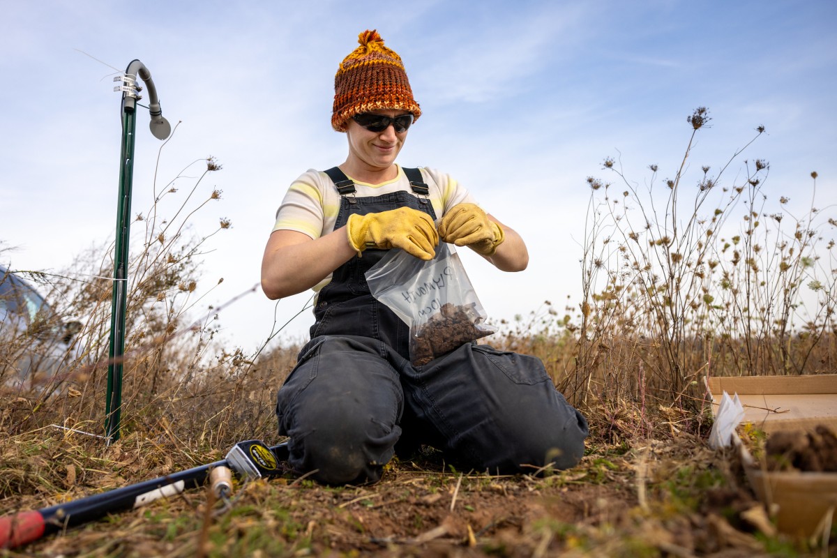 A woman in a knit cap and blue coveralls kneels on the ground sealing a clear plastic bag filled with soil.