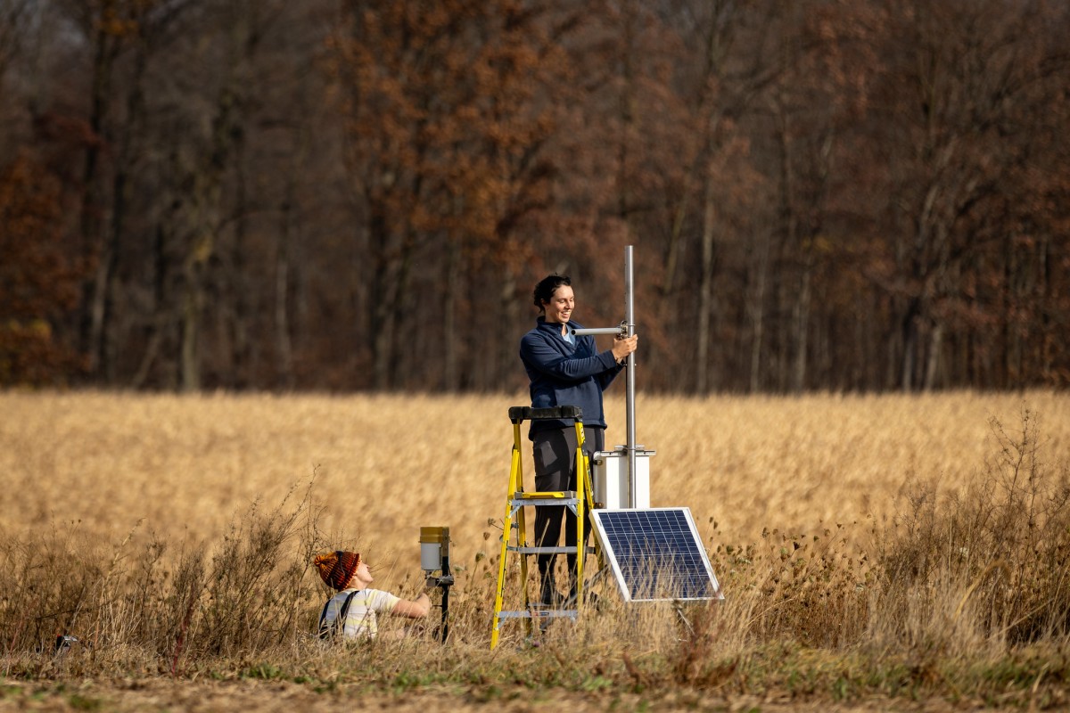 In a golden autumn field with a wooded background, two people are setting up environmental monitoring equipment. One person stands on a yellow ladder working with a metal pole while another person in a knit winter hat works at ground level. The setup includes a solar panel and scientific instruments. 