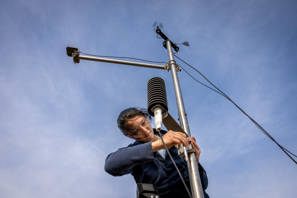 A woman attaches a piece of scientific equipment to a metal pole as seen from below. The sky in the background is blue with whisps of white clouds.