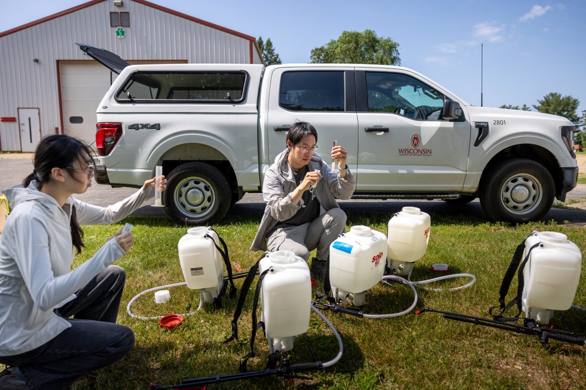 Two people crouch on the ground and inspect beakers of white fluidsurrounded by white plastic jugs. A white pickup truck is parked in the background.