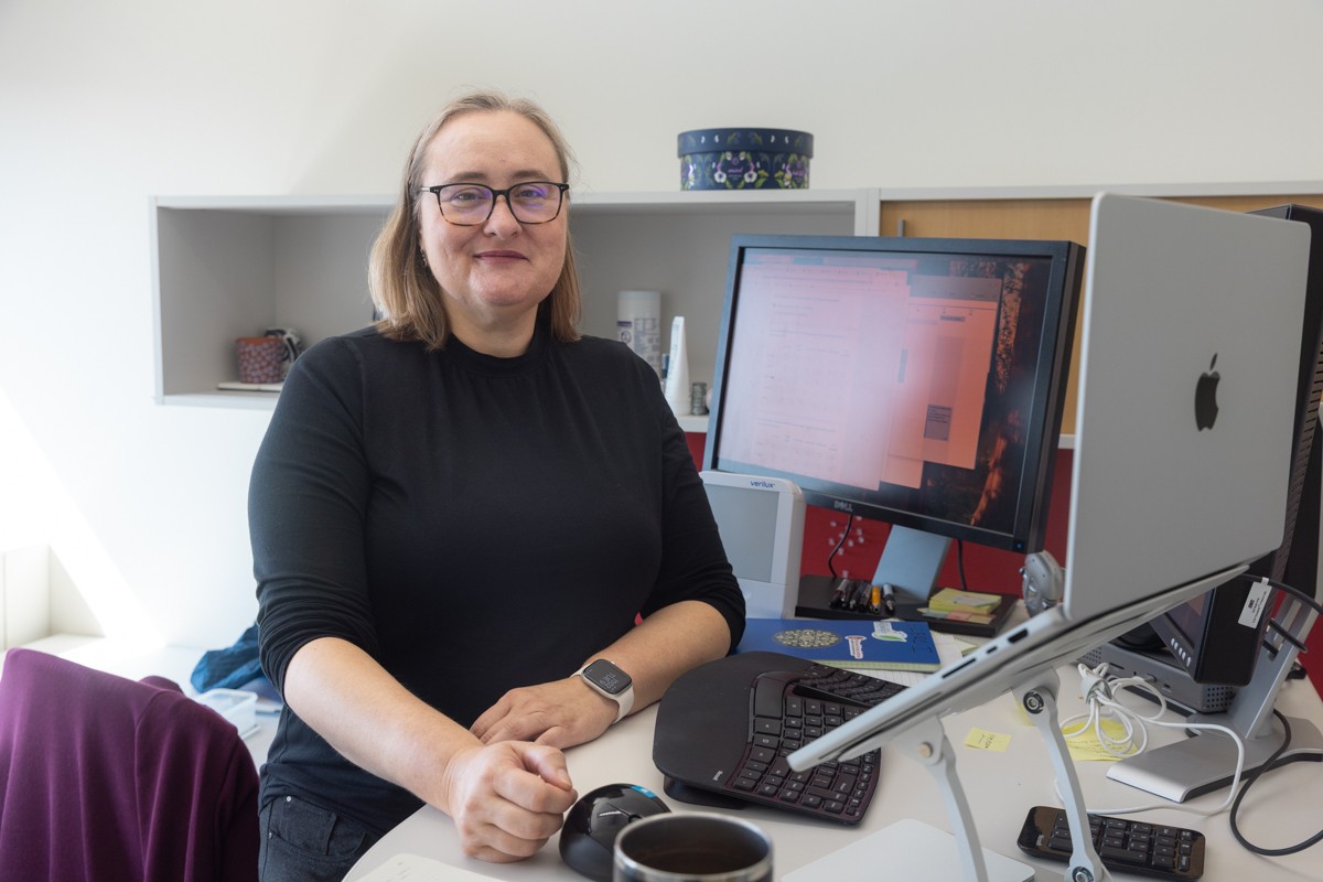 Woman with blonde hair and glasses sits at a desk with a computer monitor. She is turned to her right to face the camera