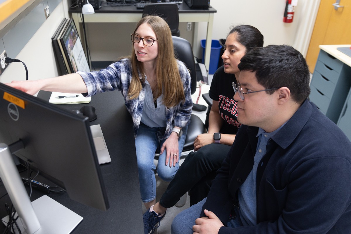 A woman points to a computer monitor as a woman and man look on
