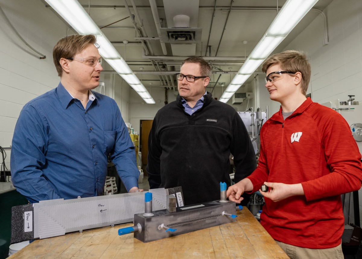 Three researchers wearing safety glasses discuss compact heat exchanger prototypes on a lab workbench, with a University of Wisconsin student in a red "W" pullover and two colleagues examining the metal test hardware.