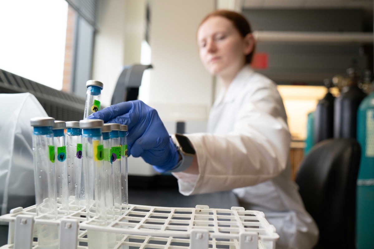Close-up of a gloved hand selecting a labeled glass vial from a test tube rack in a lab, with a blurred researcher in a white coat in the background.