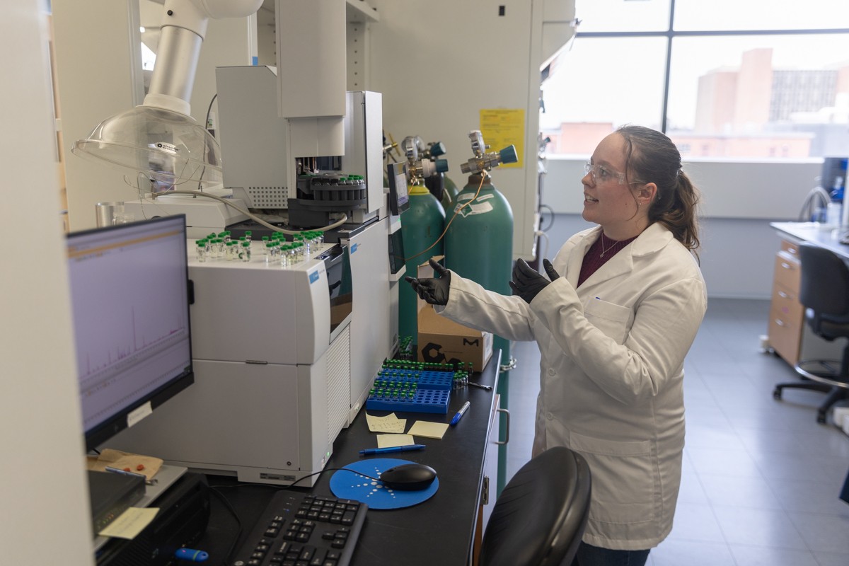 A woman in a white lab coat and glasses gestures towards a large lab machine