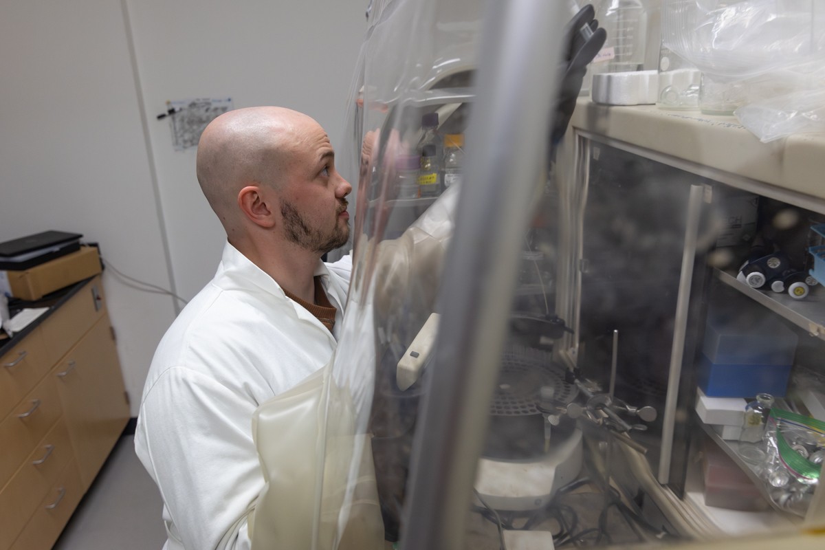 A bearded man in a white lab coat reaches for an object as he stands in front of a clear plastic barrier