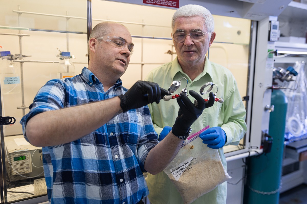 Man wearing black rubber gloves holding a piece of lab equipment as man holding plastic bag of wood chips looks on