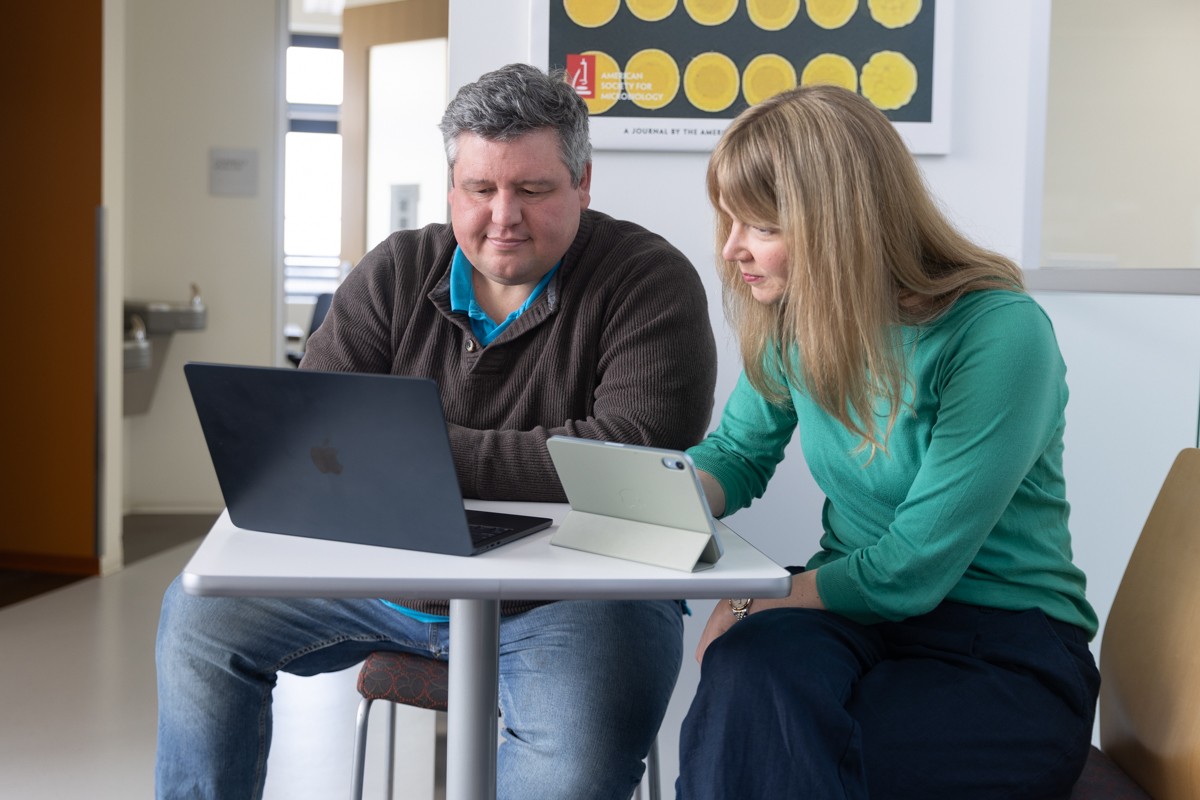 A man and woman seated at a table look at a laptop computer