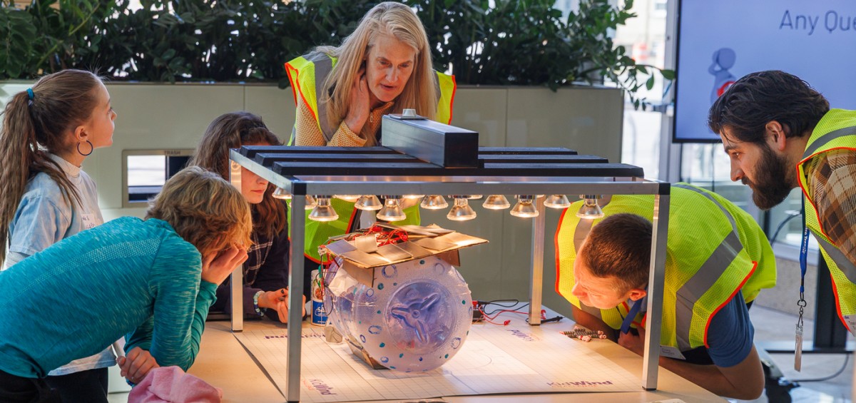 People stand around a table looking at a clear plastic cylinder with solar panels attached sitting under a bank of bright lights 