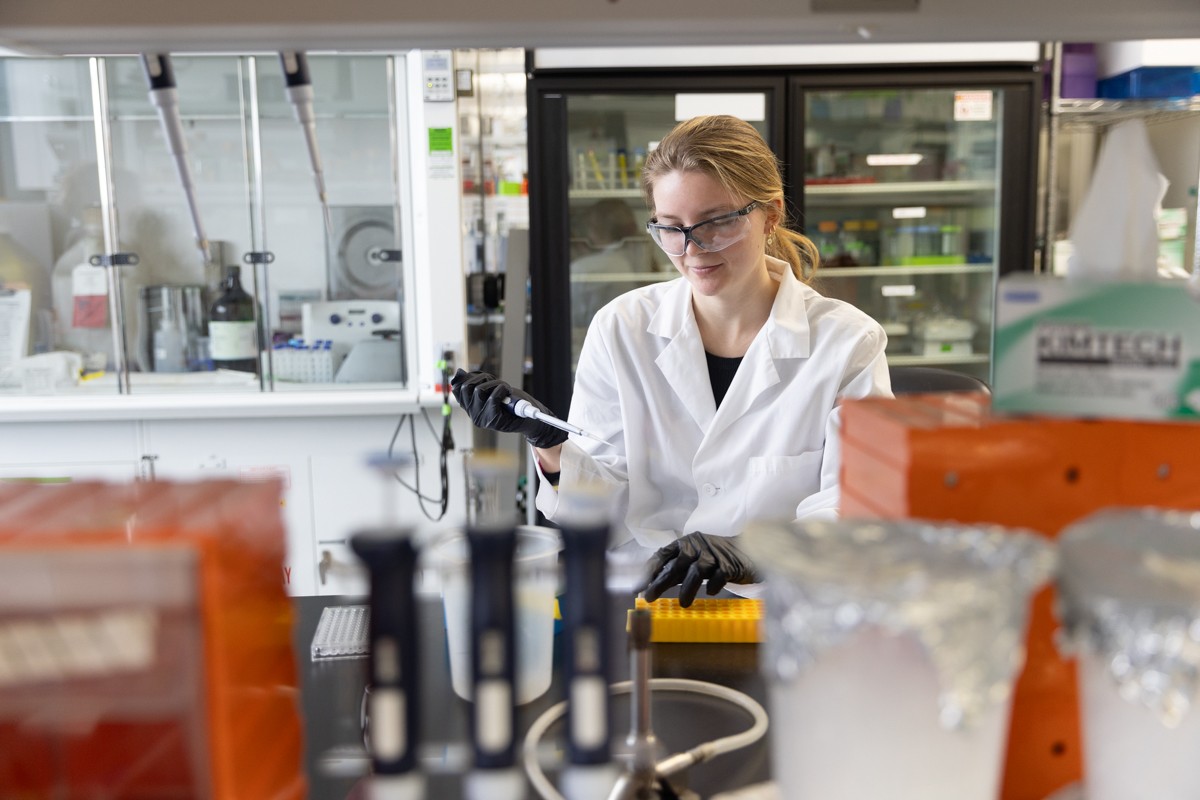A woman in a white lab coat and lab glasses holds a pipette