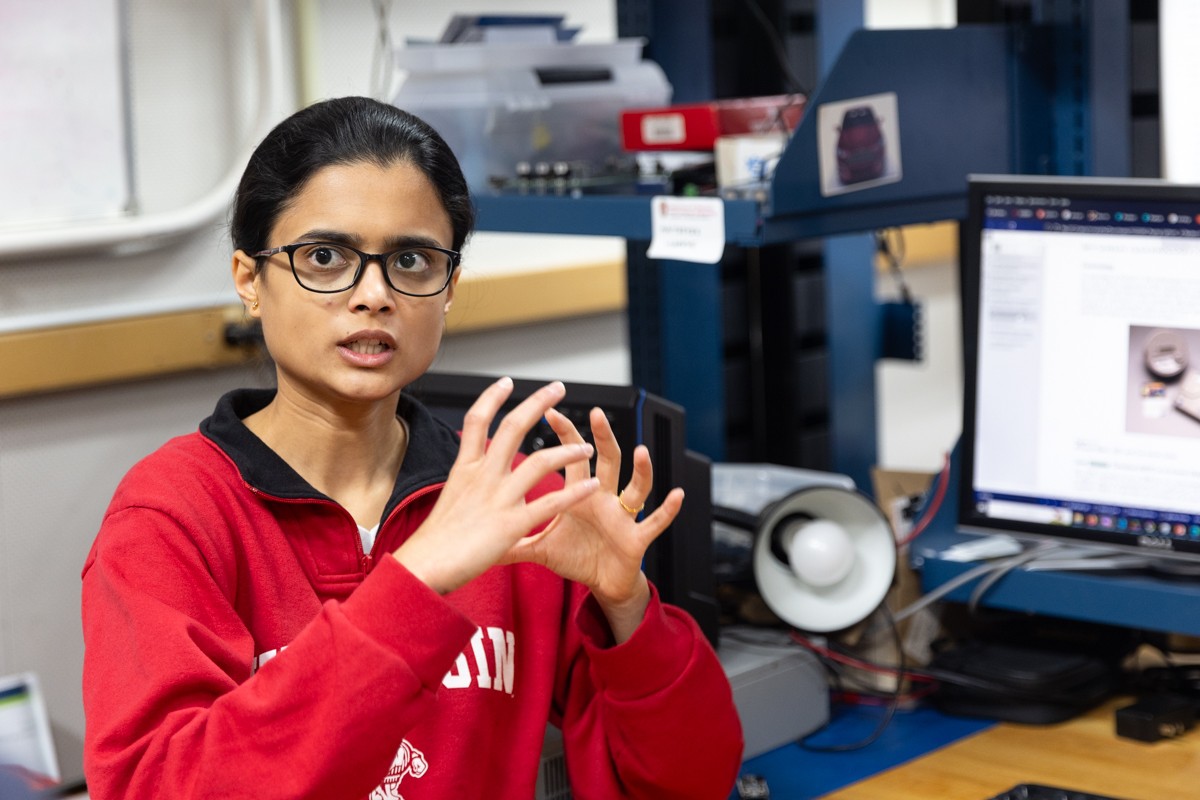 Woman with black hair and glasses in a red shirt gestures with her hands in front of a workbench with computer monitors and electronic equipment