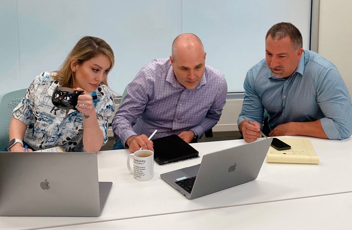A woman and two men seated at a table look at a laptop computer screen