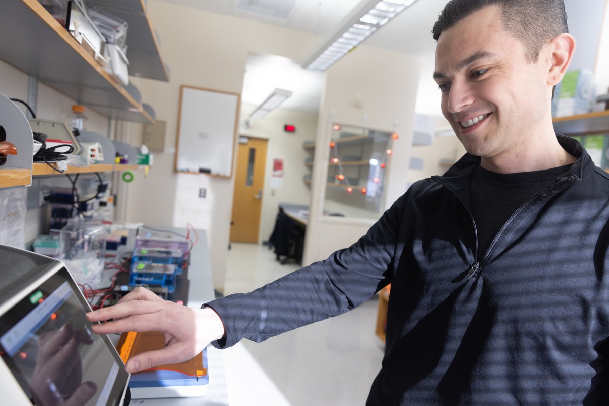 A man smiles while operating a touchscreen gel electrophoresis instrument in a lab, with shelves of lab equipment and a whiteboard visible in the background
