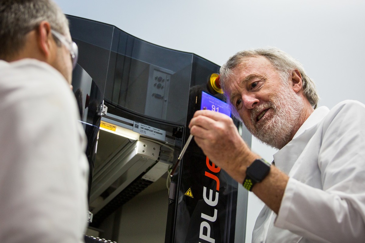 Two researchers in white lab coats examine a sample being loaded into an automated NMR or chromatography instrument (SampleJet autosampler), with a digital display showing sample number 91.
