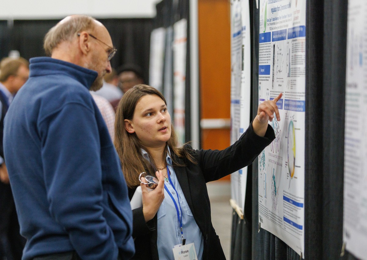 A woman wearing a name badge reading 'Rose' points to a data figure on a scientific poster while explaining it to a man in a blue fleece, at a poster session with other attendees visible in the background.