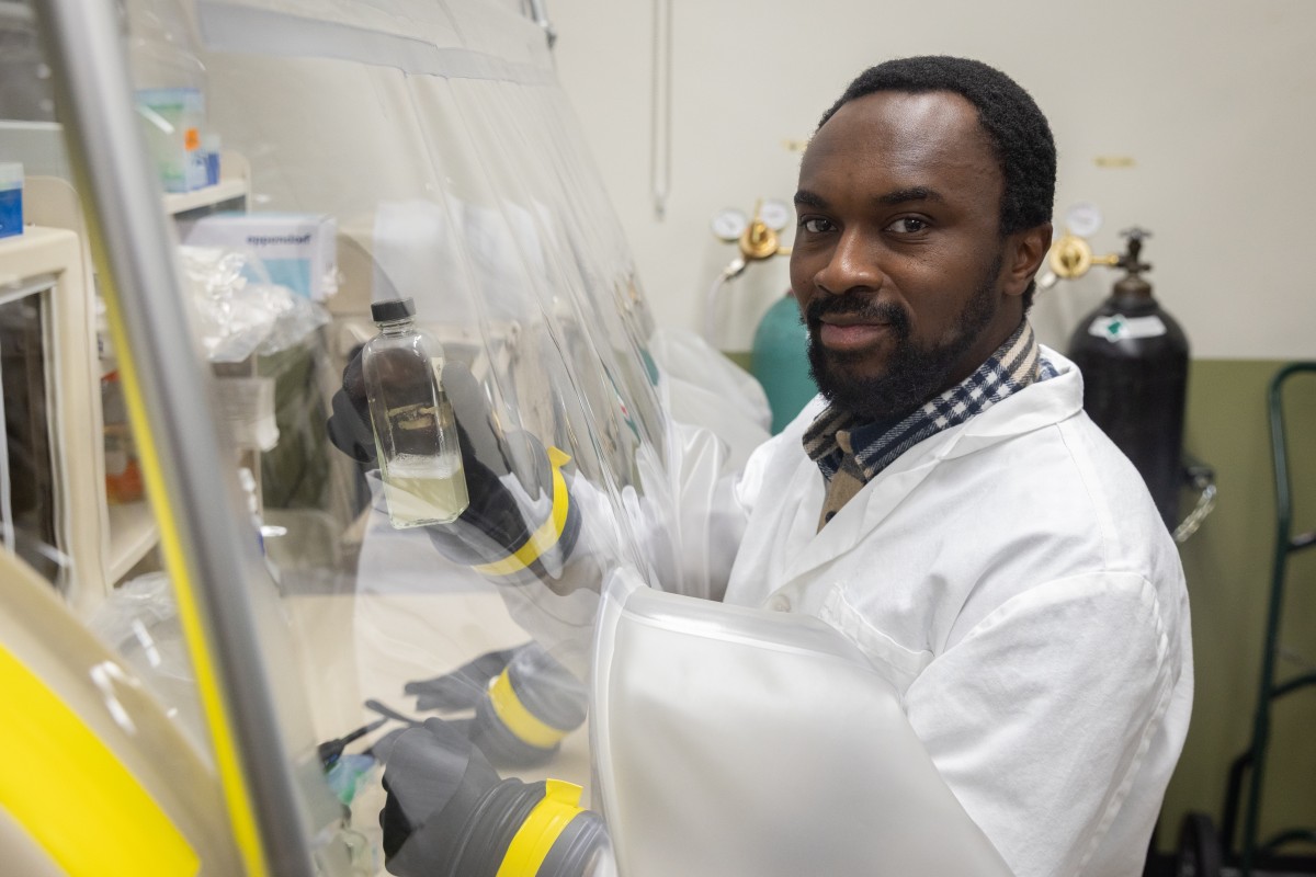 A man in a white lab coat holds a tube full of opaque liquid under a plastic sheet