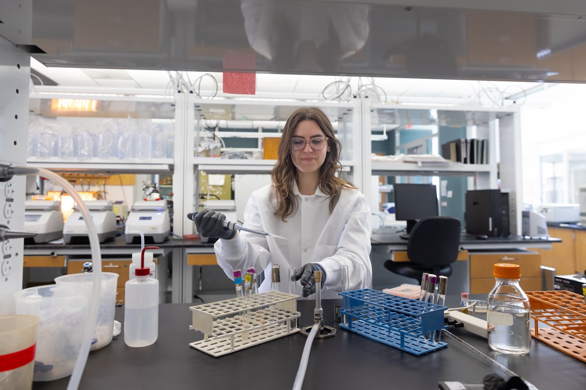 A woman in a white lab coat and latex gloves holds a flask of clear liquid in a laboratory