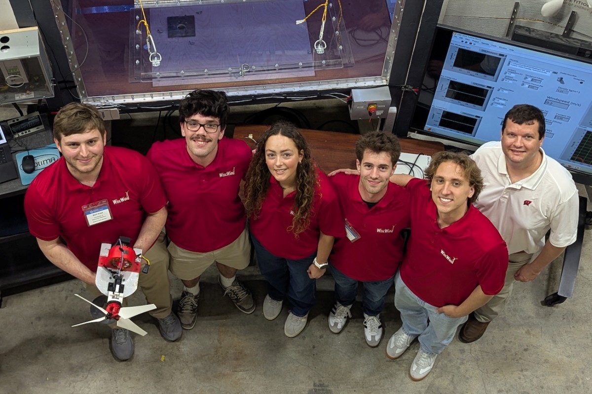 The WiscWind student team in matching red polos poses in front of a wind tunnel and data acquisition monitors, with one member holding their small-scale wind turbine prototype.