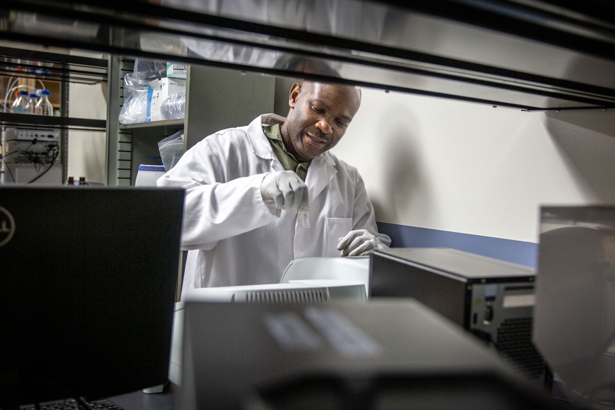 Man in white lab coat framed by dark shelving holds a glass vial